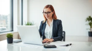 Professional woman in business attire confidently using a laptop in a bright, modern office environment with financial documents and a calculator visible on the desk